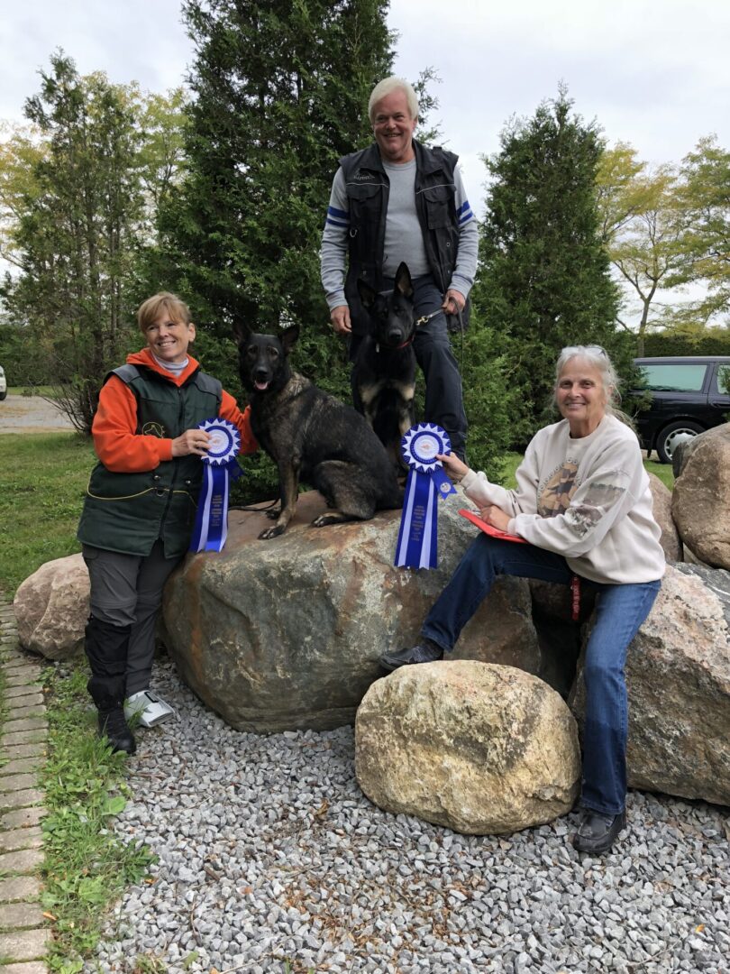 Three people holding blue ribbons stand near large boulders outdoors.