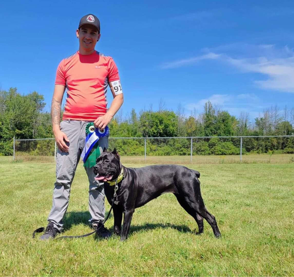 Man in red shirt standing with a black dog on a leash in a grassy field.