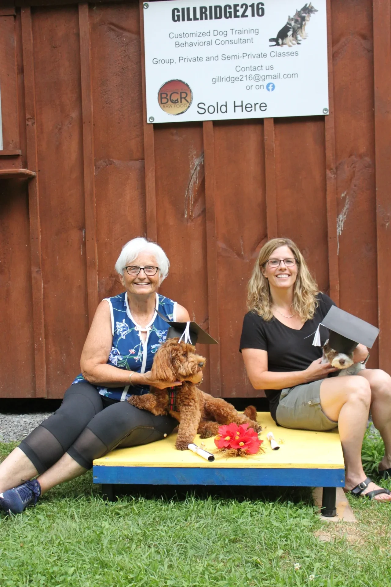 Two women sitting on a bench with a brown dog and books, smiling outdoors.